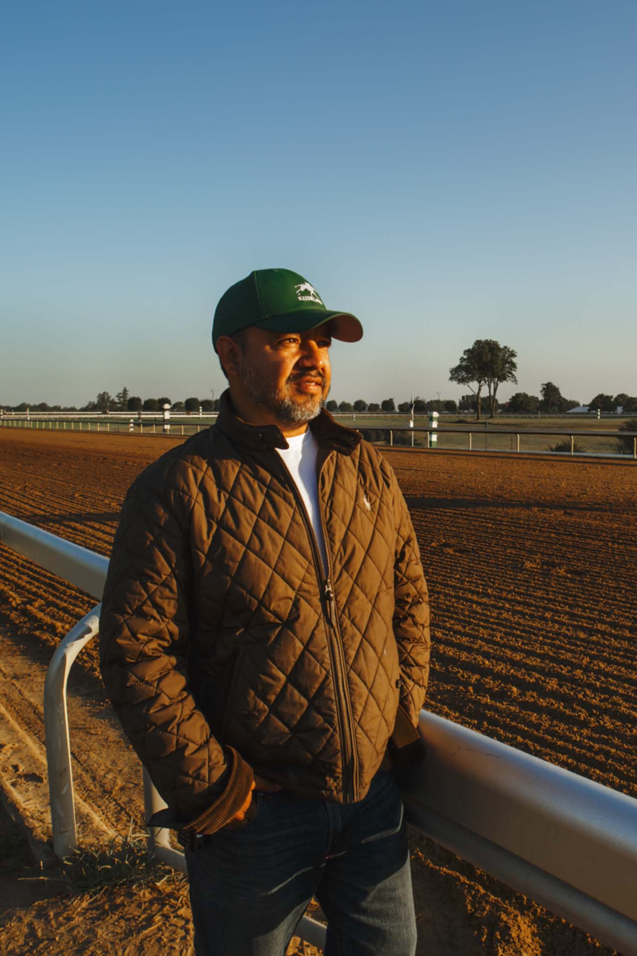 A photo of Alfredo Laureano looking off into the distance while standing by the dirt track at sunset. He is an older Hispanic man with graying chin stubble. He is wearing a green Keenfields-brand hat and a brown Keenfields-brand jacket overtop a white shirt.
