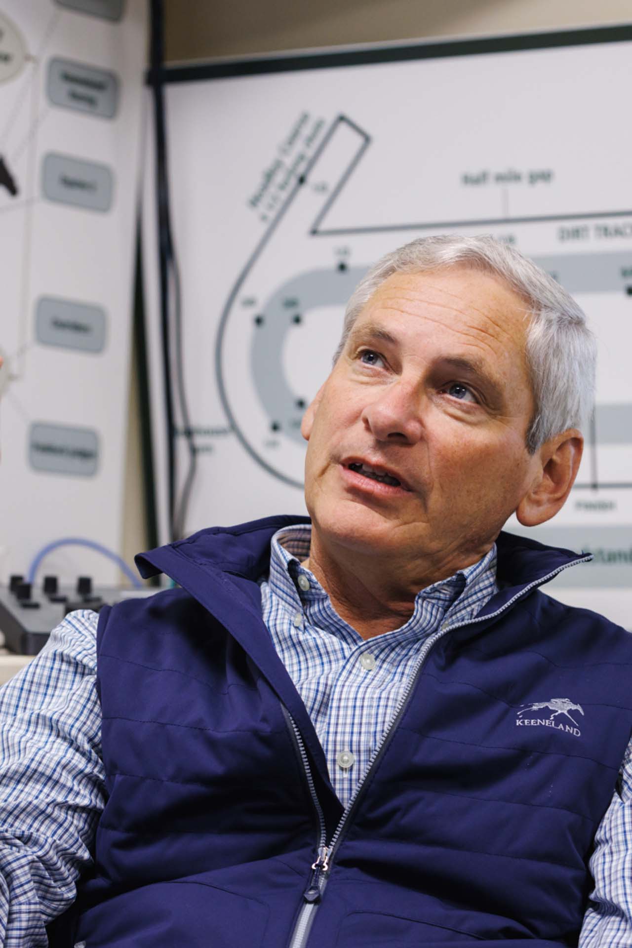 An up-close photo of Dr. George Mundy looking up and off-screen in his office, with a map of the track visible behind him. He is an older White man with short silver hair. He is wearing a navy Keenfields-brand vest over a white and blue checkered shirt.